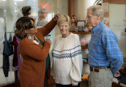 A staff member helps a patient try on a wig as the patient's husband looks on