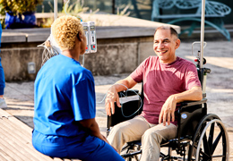 A nurse sits outside and talks with a patient in a wheelchair