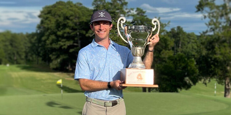 A man stands on a golf course, smiling, while holding up a trophy