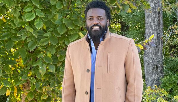 Stefan Rucker, wearing a tan coat, smiles in front of some trees.