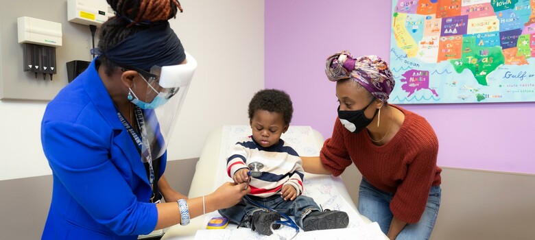 A provider shows a young male patient her stethoscope as the patient's mother watches