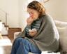 A woman sits on a couch in a blanket holding tissues. She is coughing into her hand.