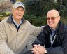 Two men sit outside at a table, smiling at the camera