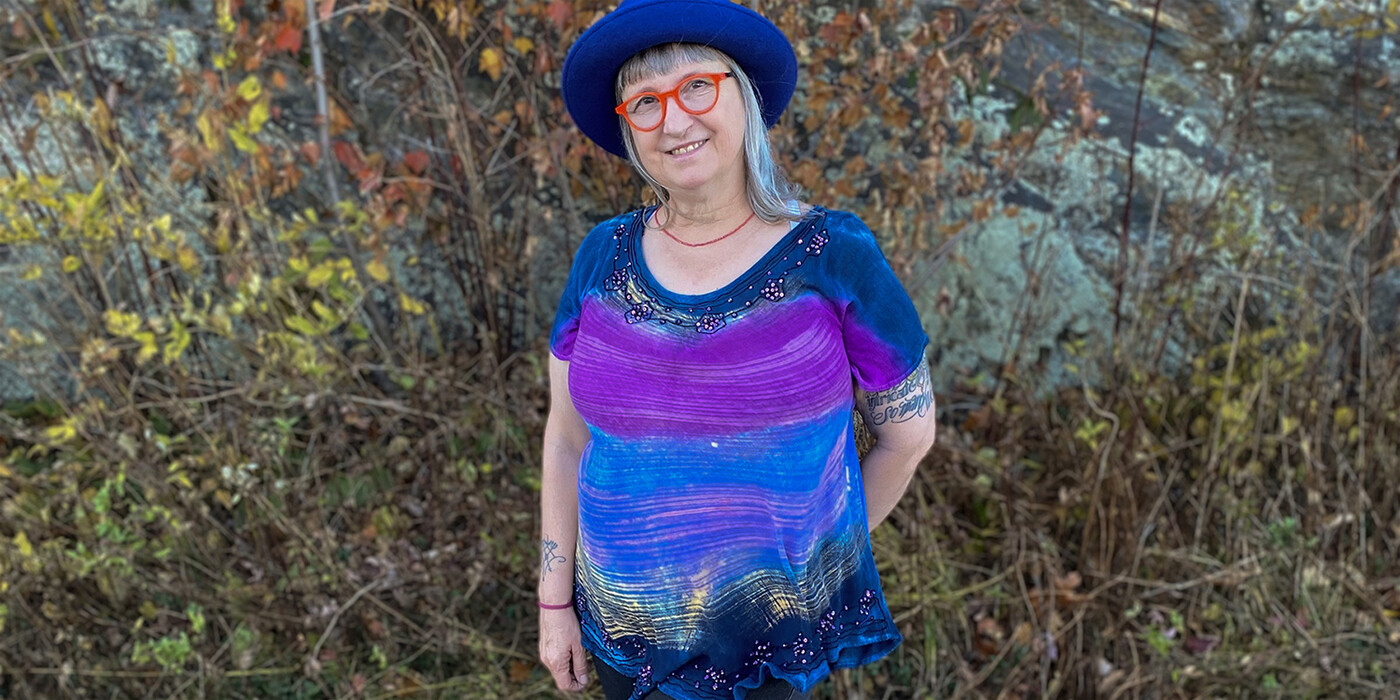 Beth Huber stands in front of some greenery. She's wearing a blue hat, orange glasses, and a blue and purple shirt.