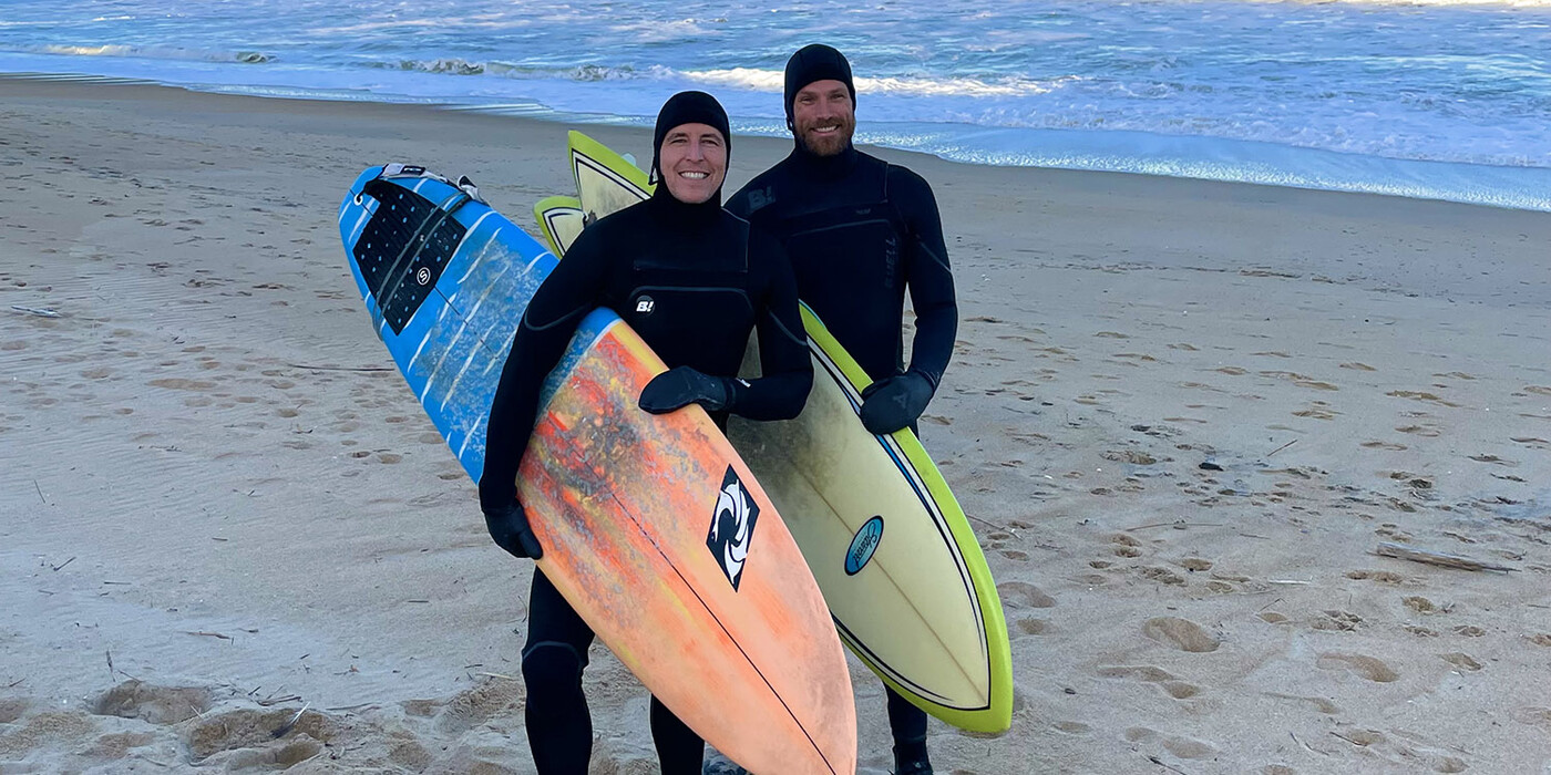 Zach Stroud and Josh Jones stand holding surfboards at the beach