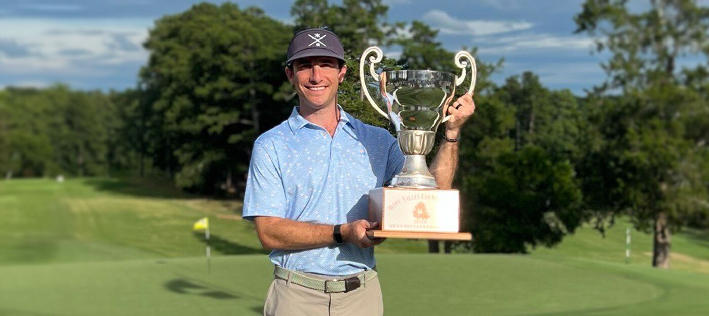 A man stands on a golf course, smiling, while holding up a trophy