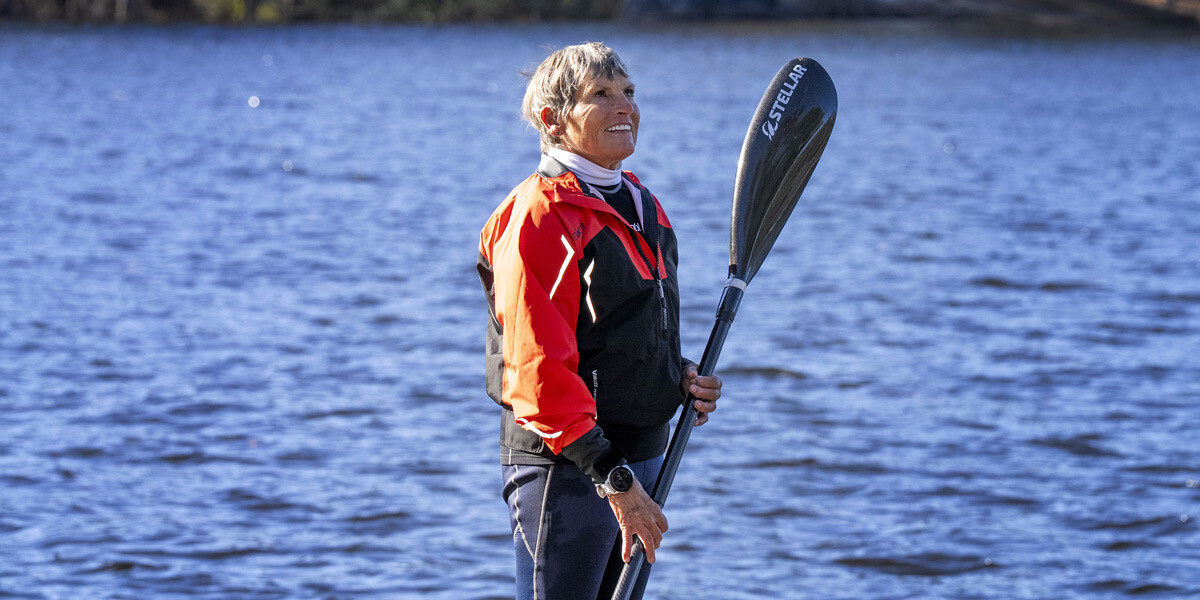 Vicki Reynolds stands in front of a lake holding her kayak padle