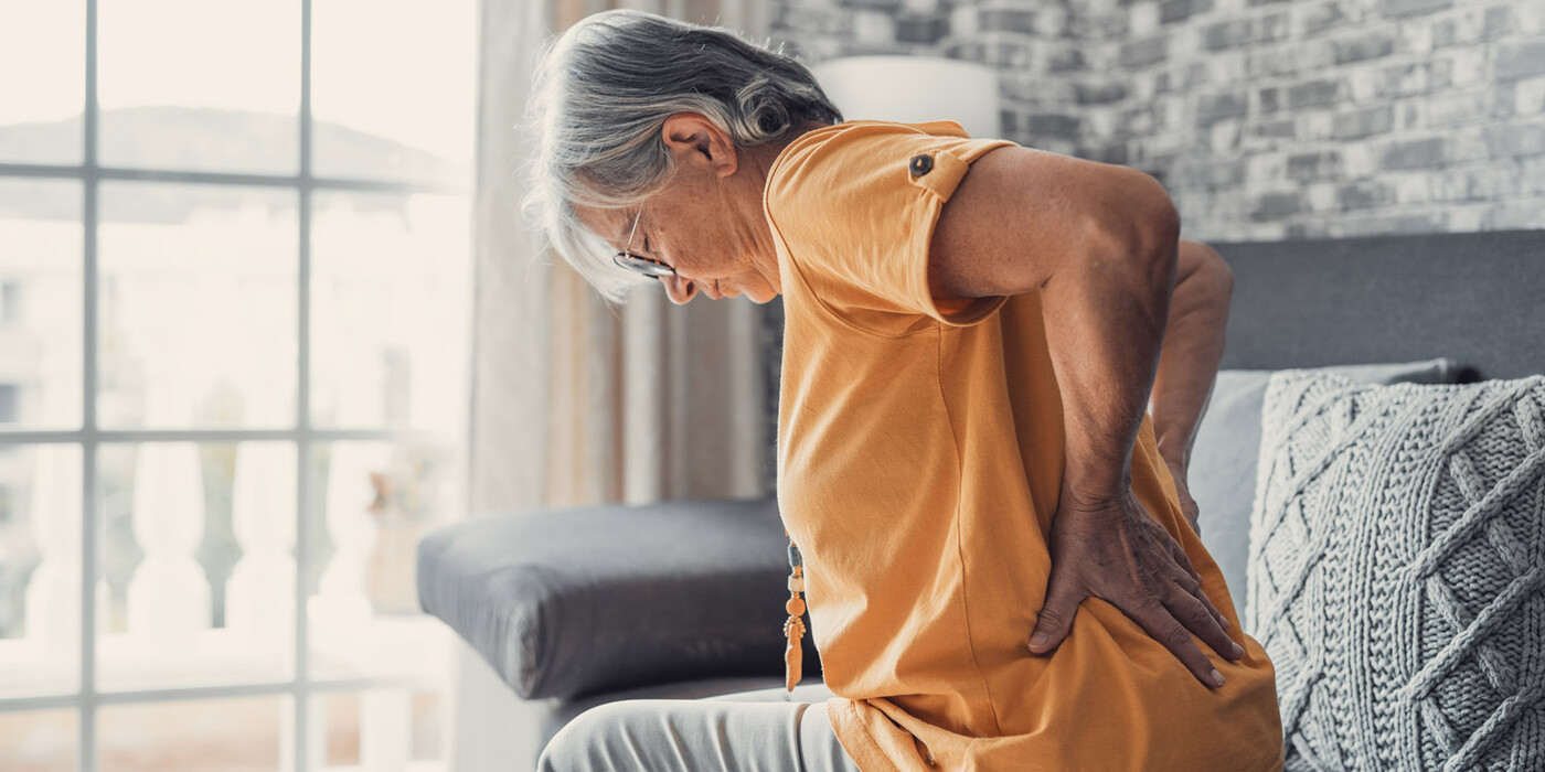 An older woman sits on a couch rubs her lower back