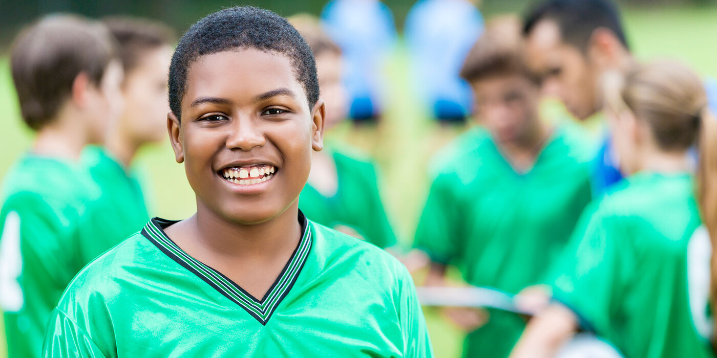 A boy wearing a soccer jersey smiles at the camera while his team huddles together behind him