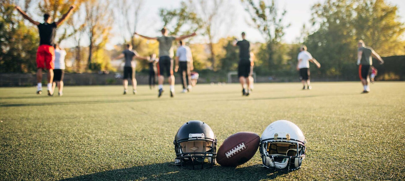Two football helmets and a football sit int he foreground as a group of players cheers in the background. 