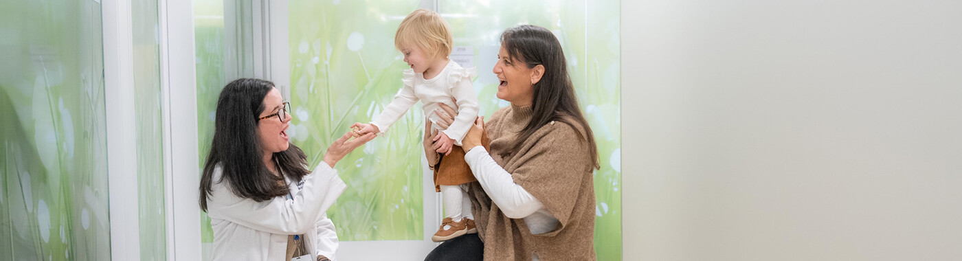 Un médico se acerca a una niña pequeña que está en brazos de su madre en un pasillo.