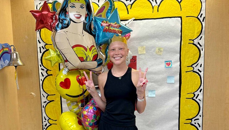 Isabella holds up peace sign fingers in front of a poster of Super Woman at Duke Children’s Hospital. 